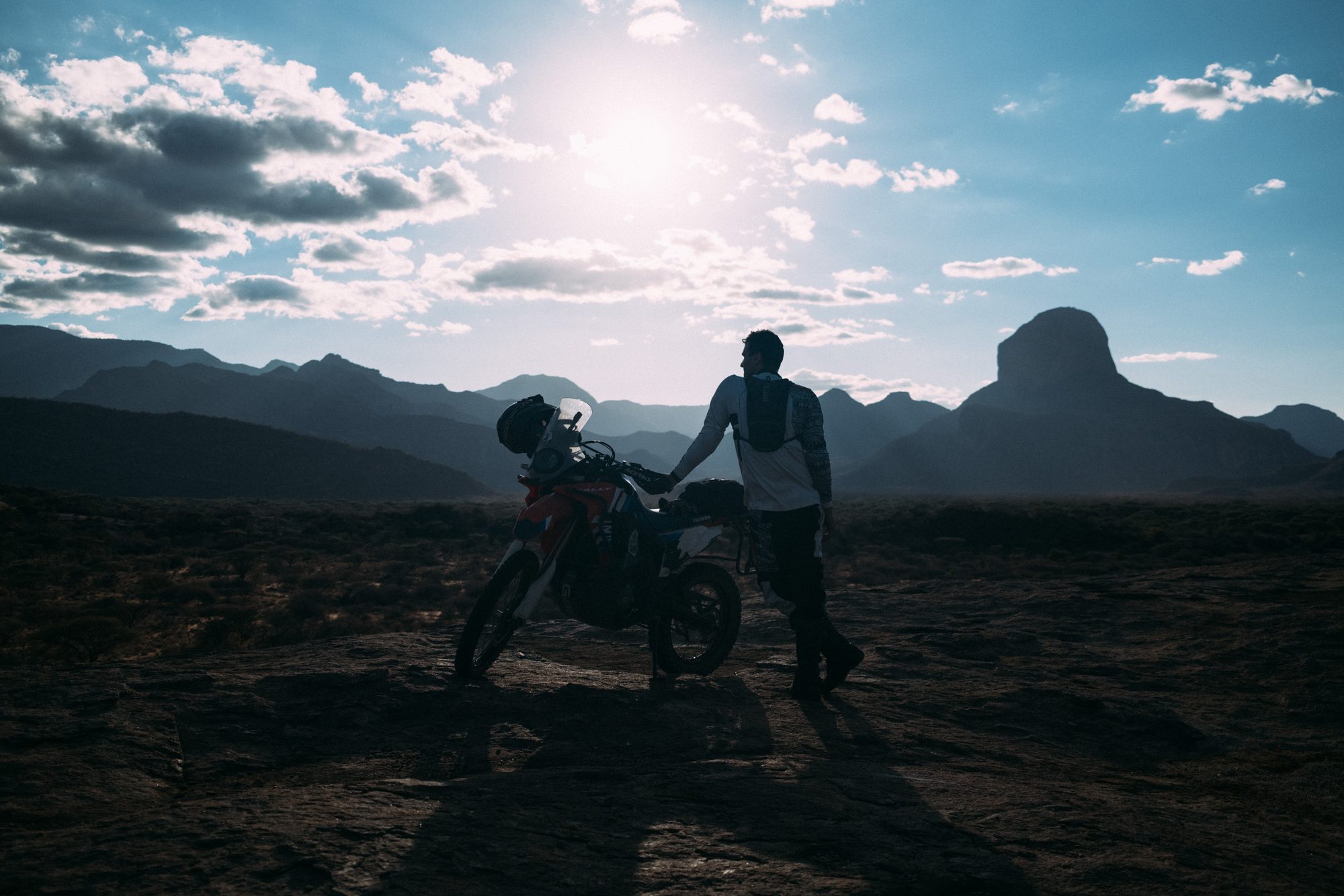 Two riders beside a motorbike on a rocky ridge at sunset, a peak behind them