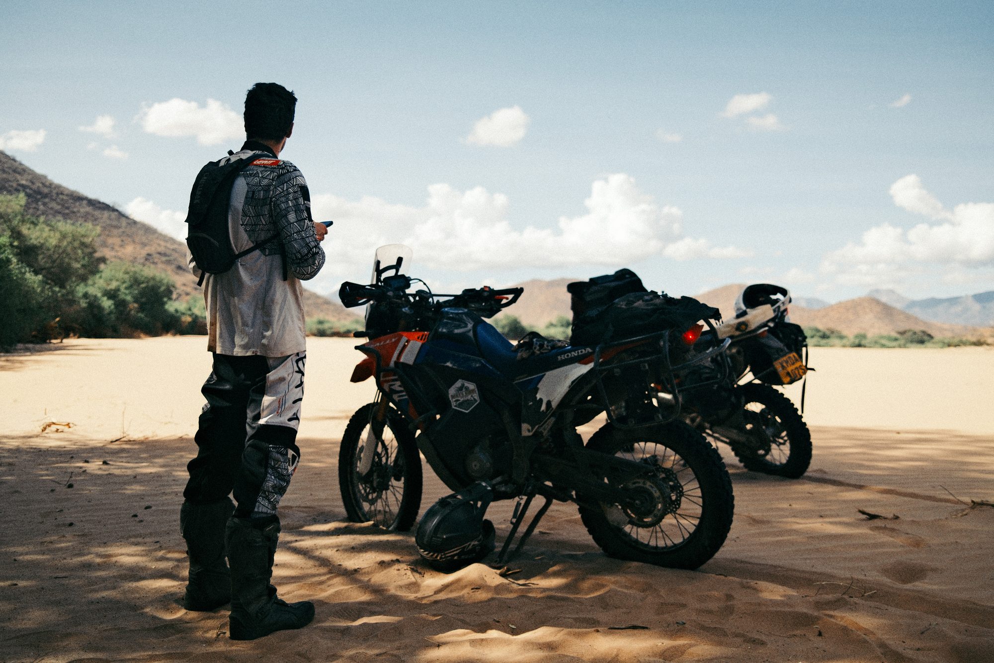 A rider standing beside two loaded bikes in the shade of a riverbank