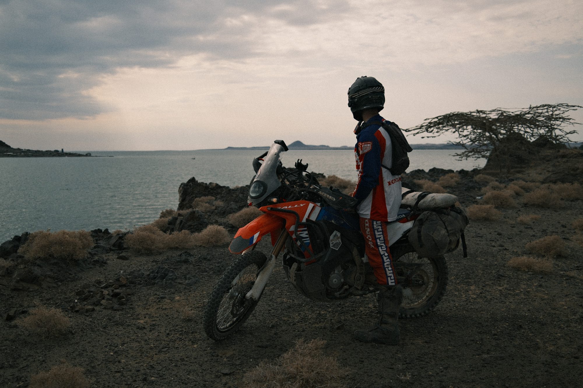 A rider standing by an orange motorbike on a rocky lakeshore